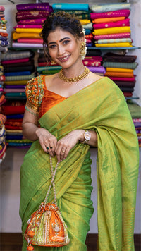 Woman in a green saree with an orange blouse, standing in front of colorful fabric shelves.