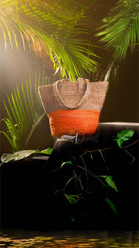 Woven bag with orange bottom half on a rock surrounded by green foliage
