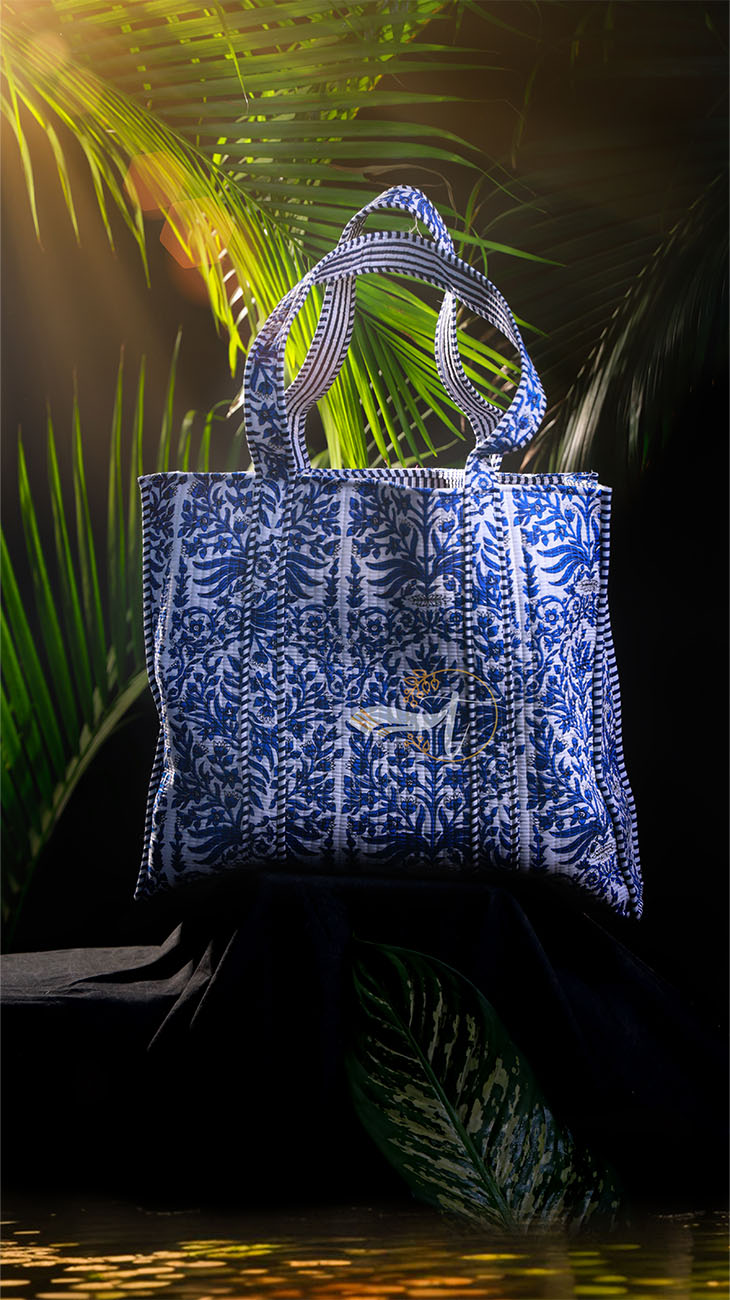 Blue patterned tote bag with a brand logo against a dark background with palm leaves.