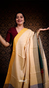 Woman in a maroon blouse and beige saree with a gold border against a dark patterned wall.