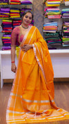 Woman in a yellow saree standing in a fabric store with colorful sarees in the background.