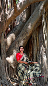 Woman in a red top and black and white checkered saree sitting under a large tree with twisted roots.