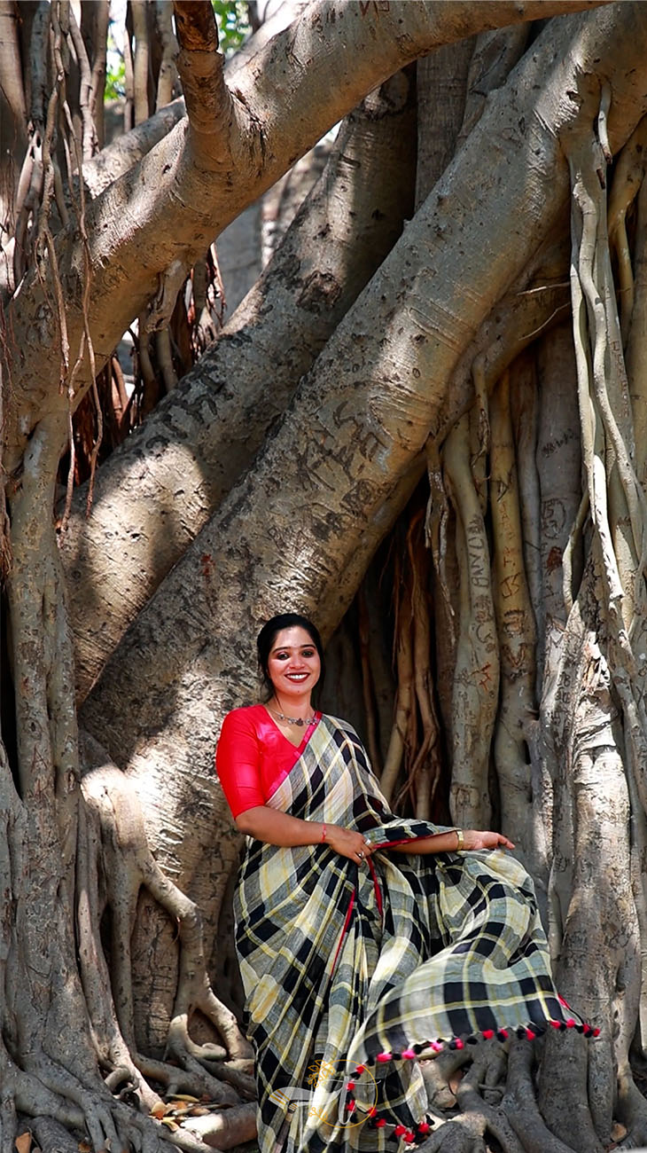 Woman in a red top and black and white checkered saree sitting under a large tree with twisted roots.