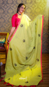 Woman wearing a bright yellow saree with pink border indoors against a patterned wall.