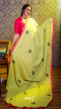 Woman wearing a bright yellow saree with pink border indoors against a patterned wall.