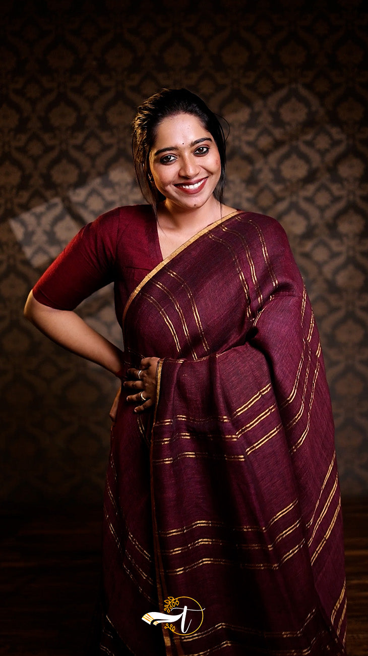 Woman in a maroon saree with gold border against a patterned wall.