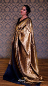Woman wearing a gold velvet saree with a blue blouse against a patterned wall.