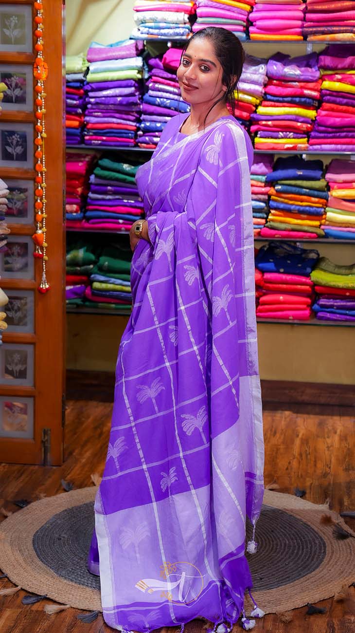 Woman wearing a purple saree with a white pattern in a store with colorful sarees in the background.