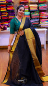 Woman in a black saree with gold border standing in front of a colorful fabric display.