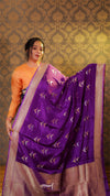 Woman wearing a purple saree with a gold border against a patterned wall.