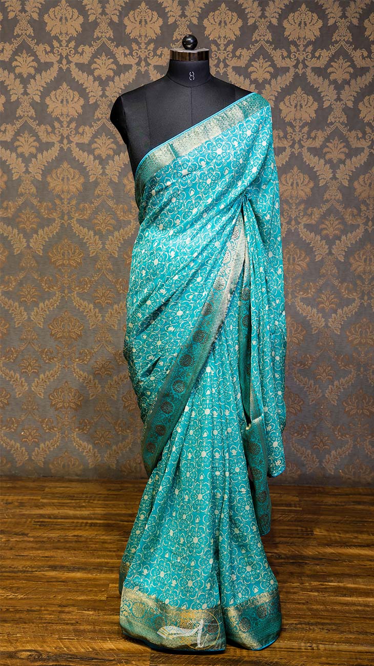 Teal saree with white patterns on a mannequin against a patterned wall.