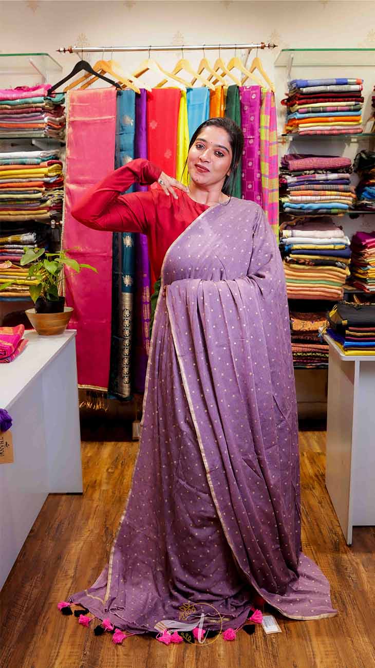 Woman in a purple saree with pink blouse standing in a store with colorful sarees in the background.
