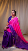 Woman wearing a pink and purple saree sitting against a plain background