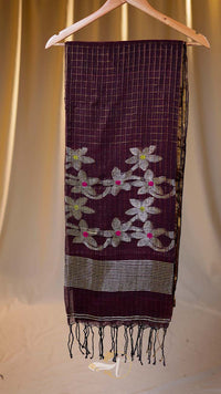 Dark purple scarf with floral patterns and fringes on a wooden hanger against a beige curtain background.
