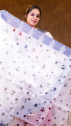 Woman wearing a white saree with floral patterns against a brown background