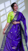 Woman wearing a purple saree with green blouse against a white wall.