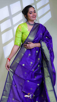 Woman wearing a purple saree with green blouse against a white wall.