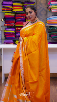 Woman in a bright orange saree standing in front of a display of colorful sarees.