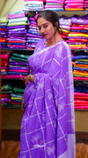 Woman wearing a purple saree with a pattern in front of colorful fabric stacks.