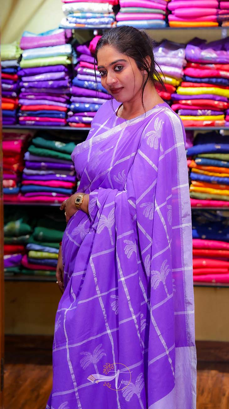 Woman wearing a purple saree with a pattern in front of colorful fabric stacks.