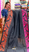 Woman holding a dark gray saree with red and pink patterns in a colorful store setting.