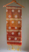 Orange scarf with white polka dots and gold fringe on a wooden hanger against a neutral background