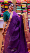 Woman wearing a purple saree with a green blouse in front of colorful sarees.