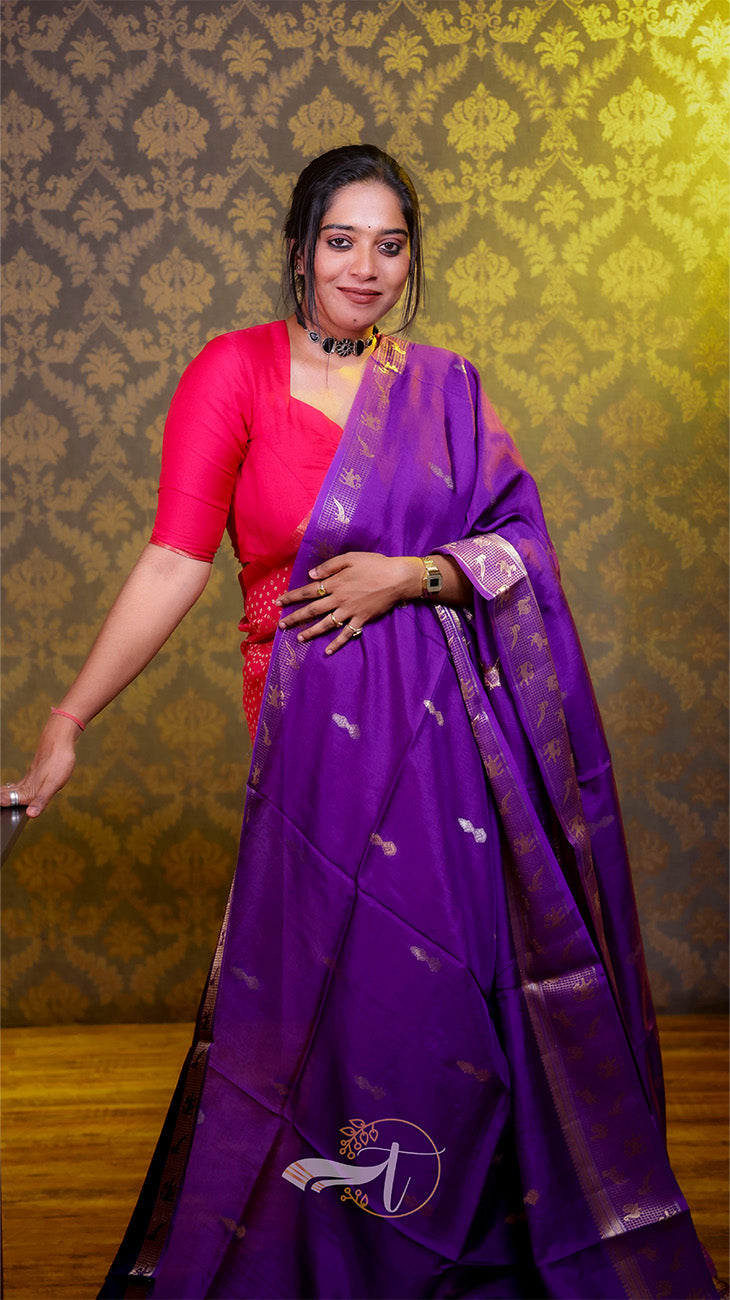 Woman wearing a purple saree with a red blouse against a patterned wall.