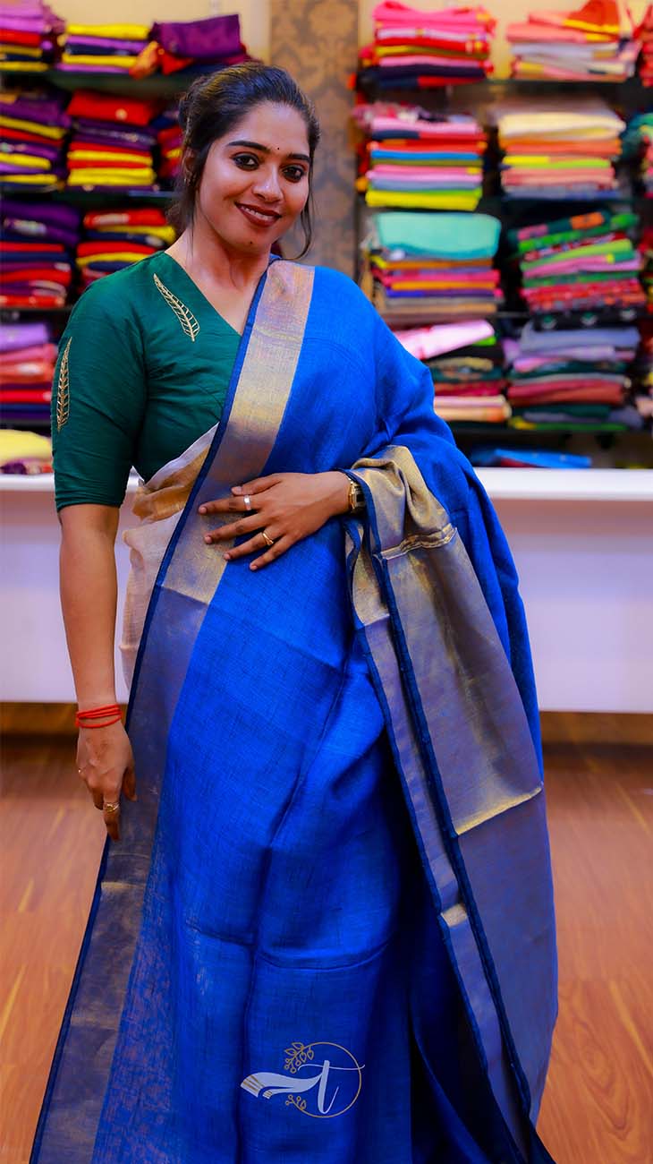 Woman wearing a blue saree with a green blouse in front of a backdrop of colorful sarees.