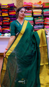 Woman wearing a green saree with yellow border in front of colorful fabric displays.