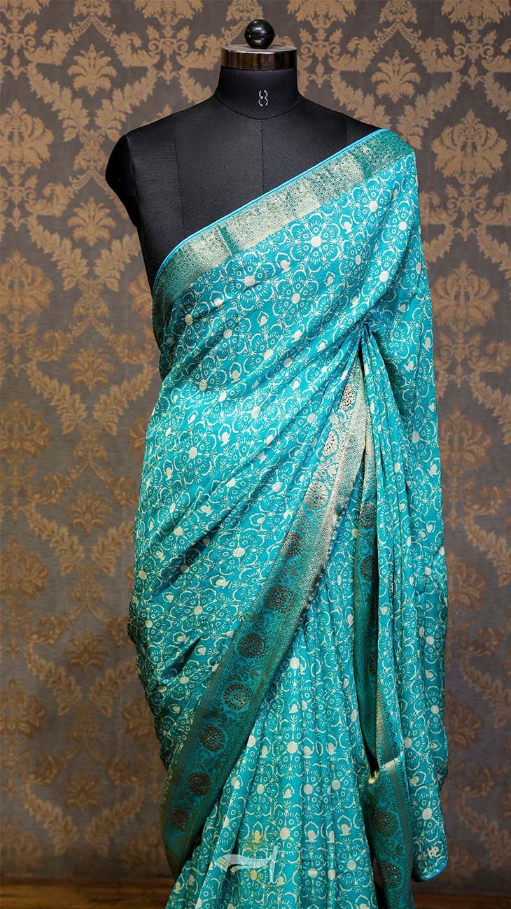 Teal saree with white patterns on a mannequin against a patterned wall.