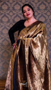 Woman wearing a gold and black saree against a patterned wall.