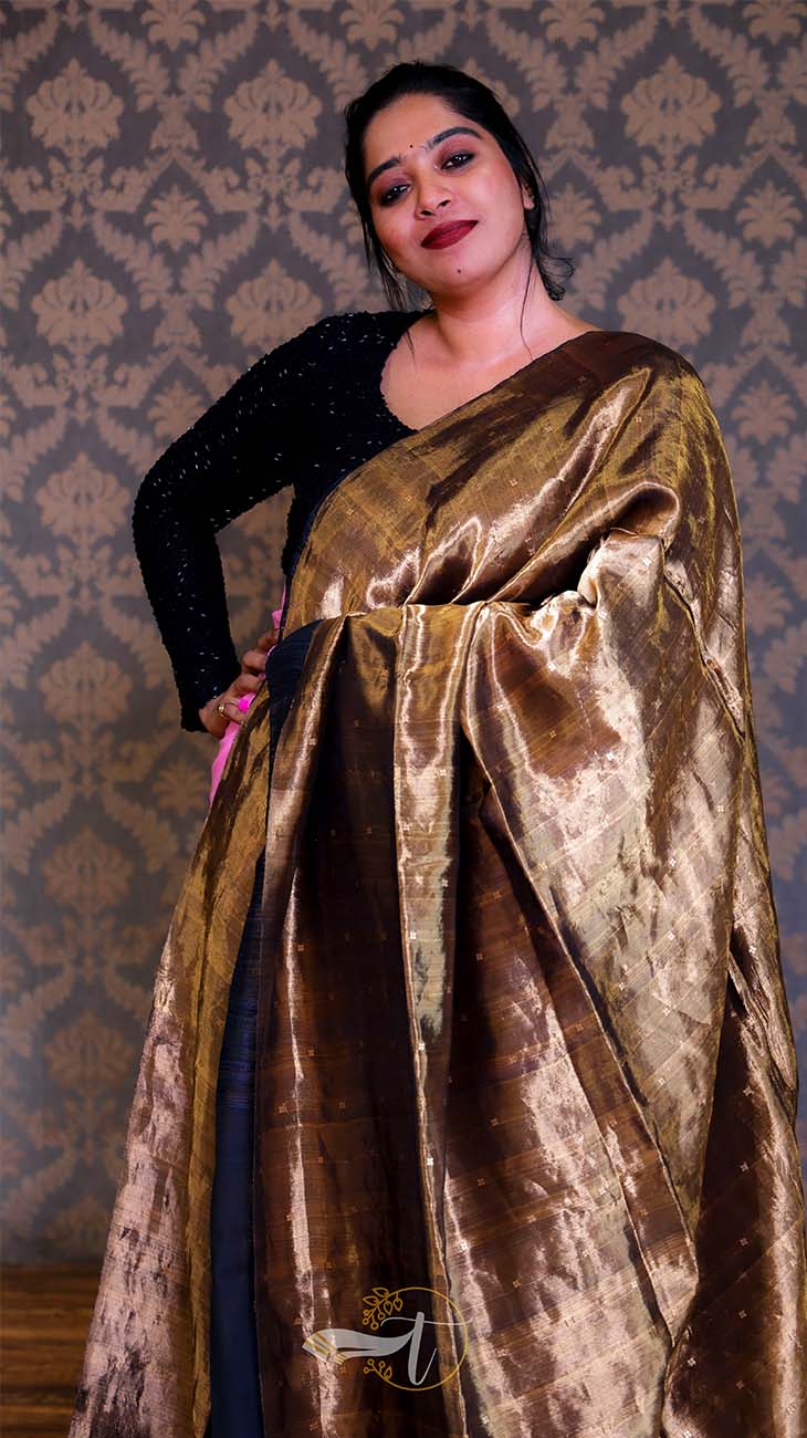 Woman wearing a gold and black saree against a patterned wall.