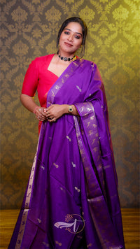 Woman wearing a purple saree with a pink blouse against a patterned wall.