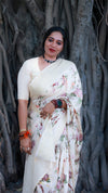 Woman in a white saree with floral patterns standing against a tree trunk.