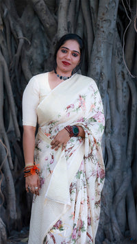 Woman in a white saree with floral patterns standing against a tree trunk.