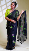 Woman wearing a black saree with green and purple floral patterns, holding a matching black dupatta.