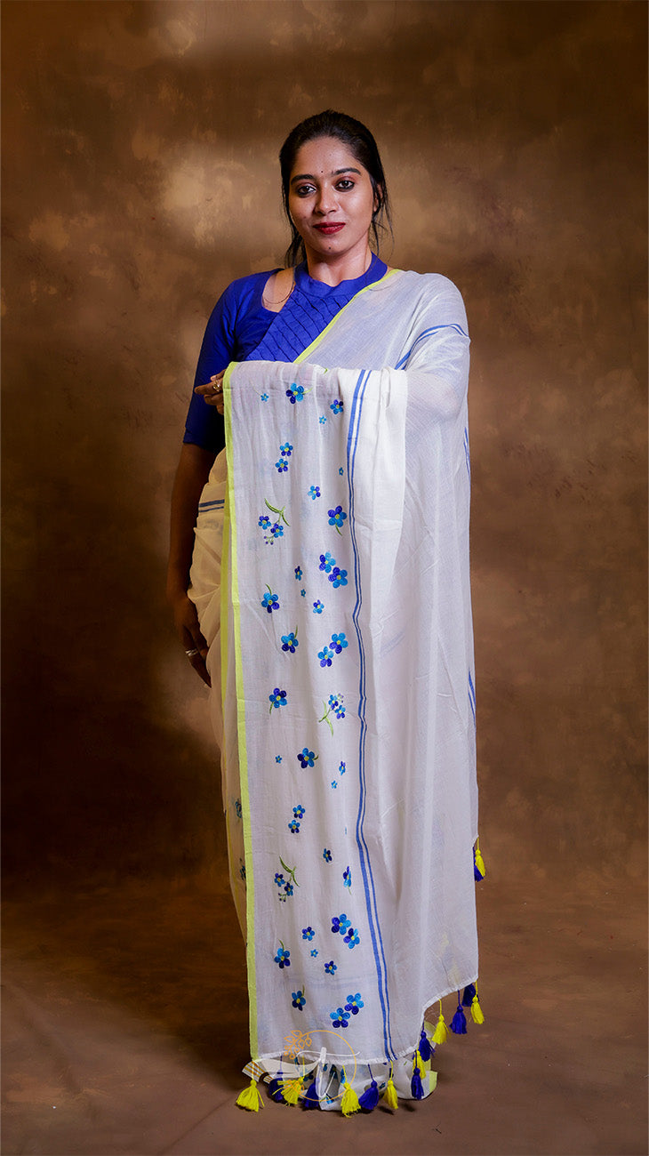 Woman wearing a white saree with blue floral patterns and yellow border against a brown background