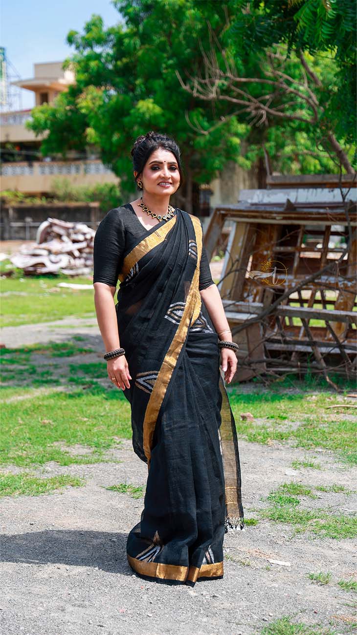 Woman in a black saree with gold border standing outdoors on a sunny day.