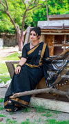 Woman in a black saree with gold border sitting outdoors near wooden structures.