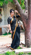 Woman in a black saree with gold border standing next to a tree outdoors.
