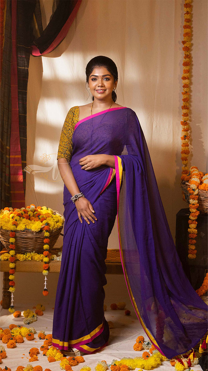Woman in a purple saree with yellow border standing in a decorated room.
