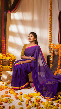 Woman in a purple saree sitting on a decorated floor with flowers and marigold garlands.