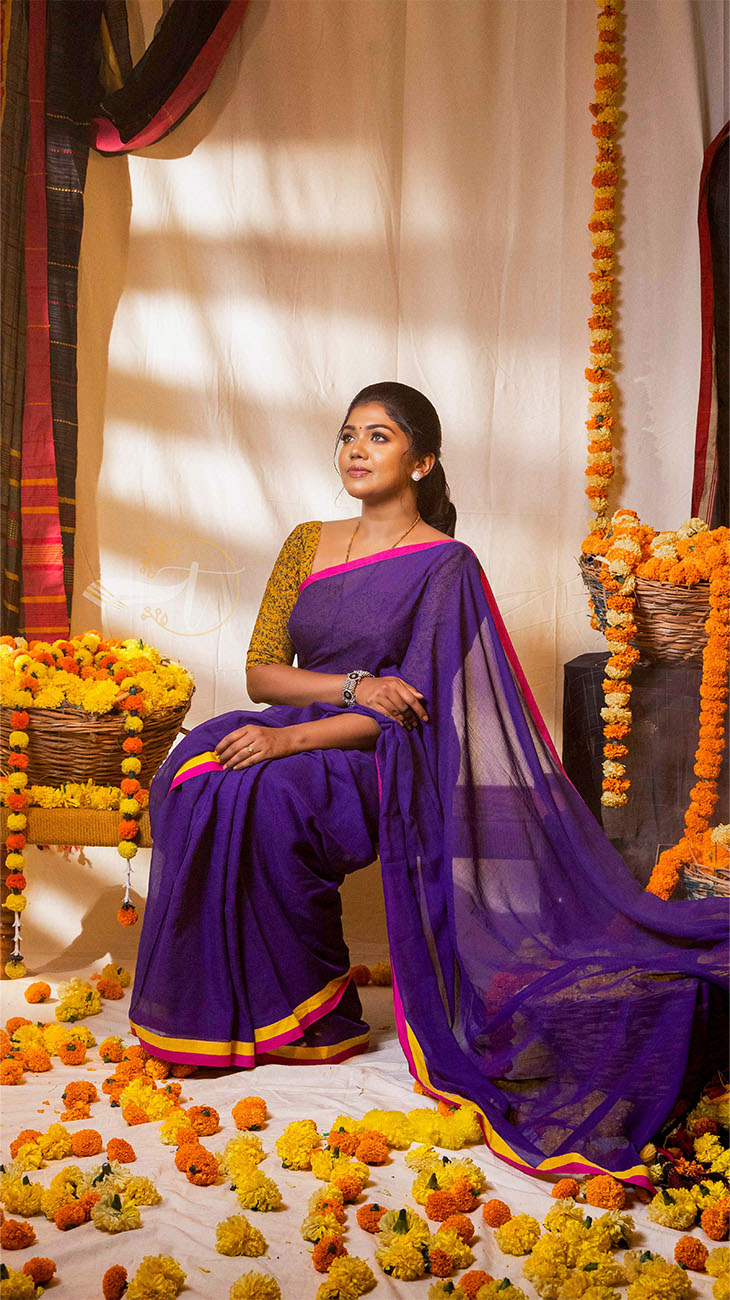 Woman in a purple saree sitting on a decorated floor with flowers and marigold garlands.