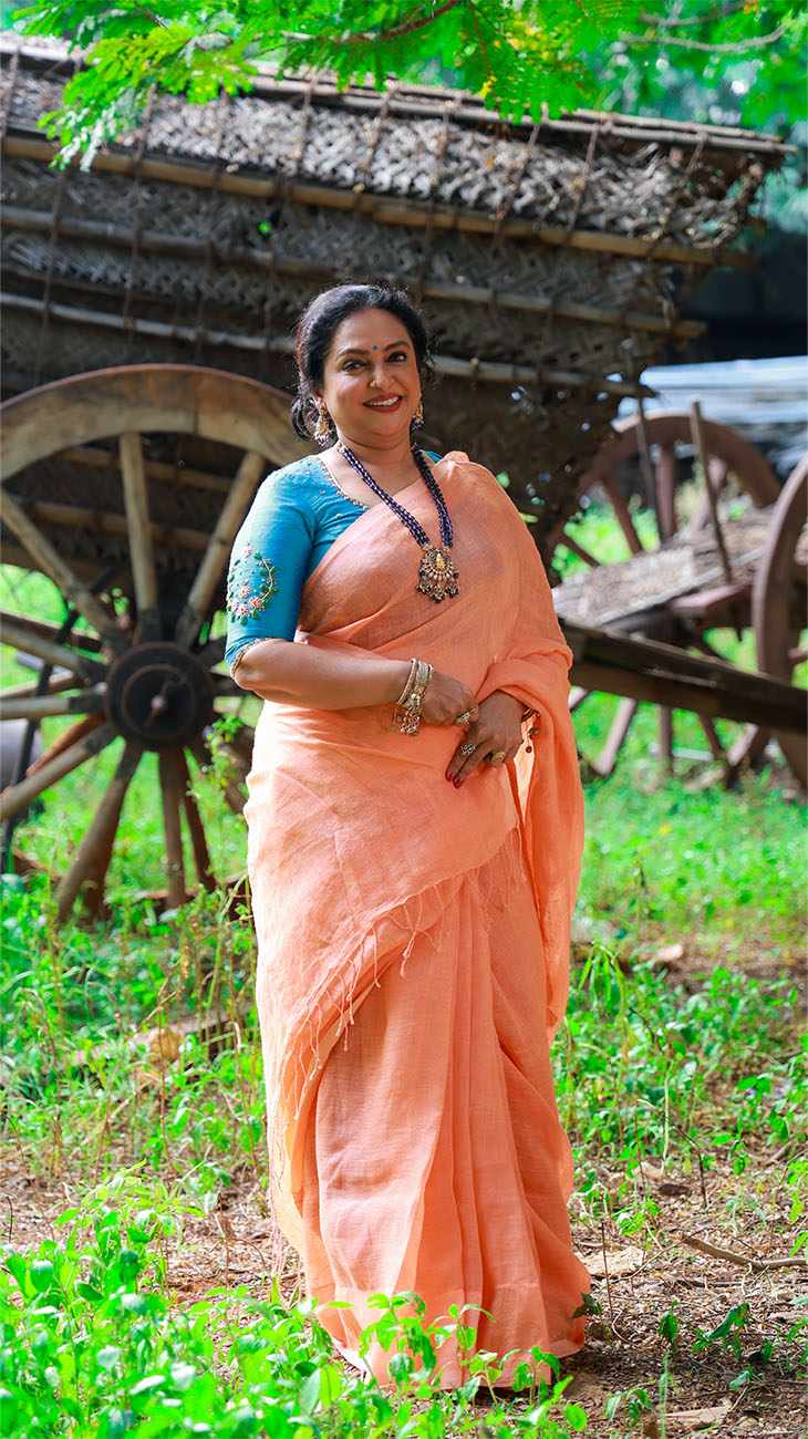 Woman in a peach saree standing in a natural setting with wooden carts in the background