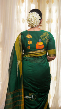 Woman wearing a green saree with intricate patterns, standing against a white curtain.