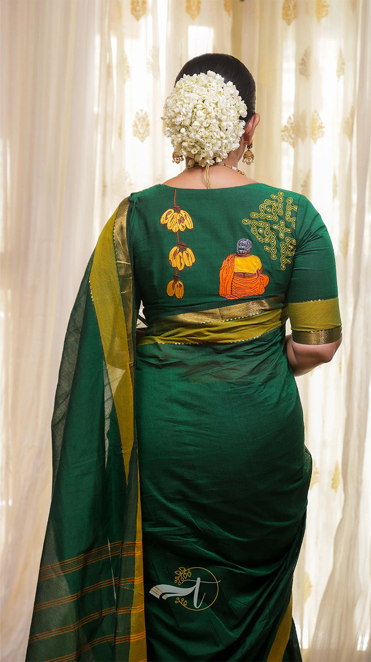 Woman wearing a green saree with intricate patterns, standing against a white curtain.