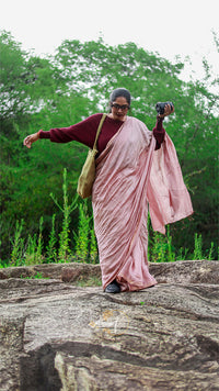 Woman in a pink saree standing on a rock with greenery in the background