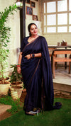 Woman in a dark blue saree standing in a room with plants and a window.