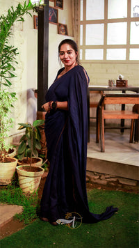 Woman in a dark blue saree standing in a room with plants and a table.
